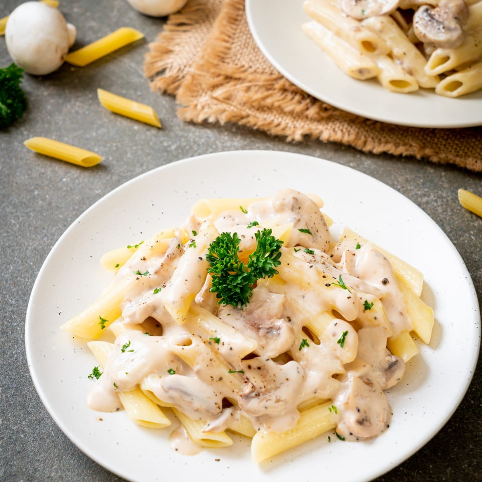 Two plates of penne pasta with creamy mushroom sauce, garnished with parsley and black pepper.