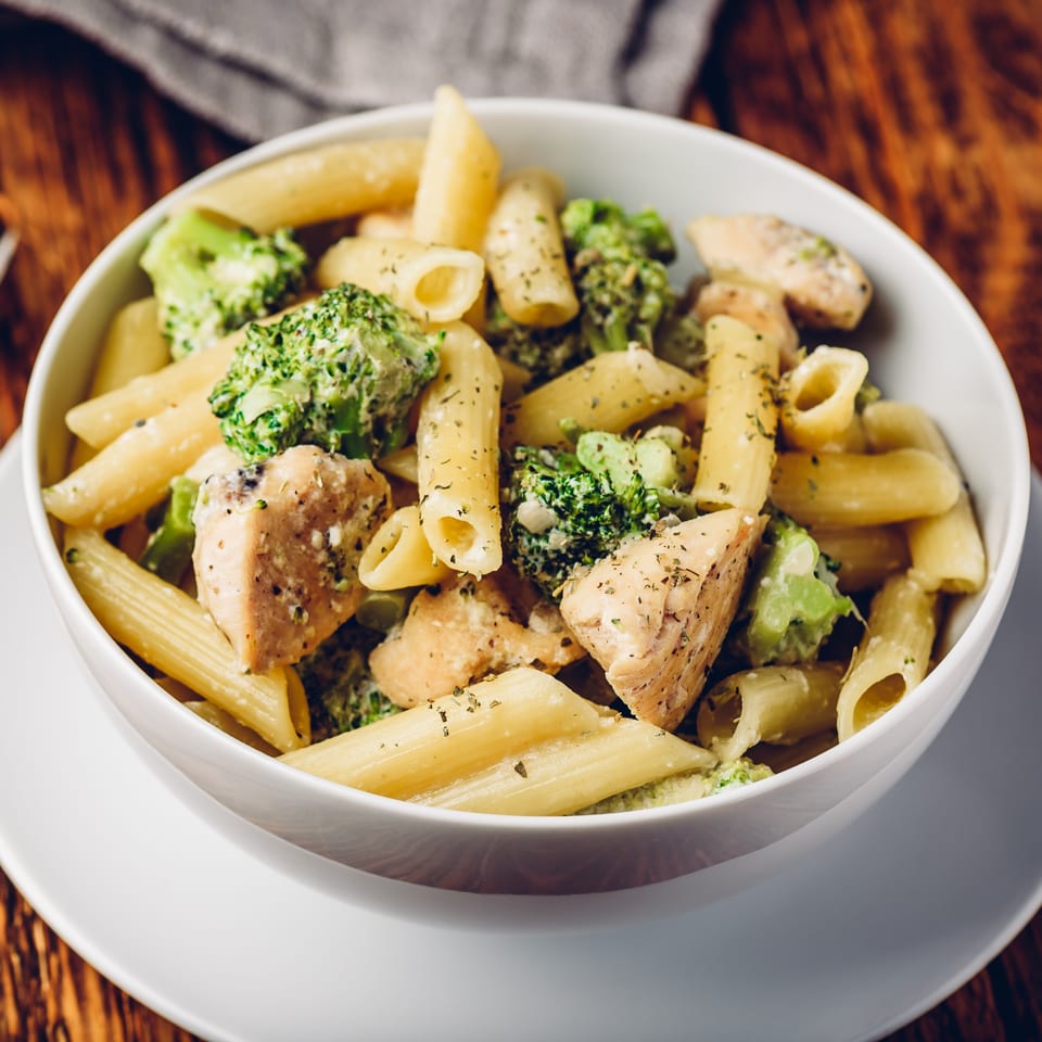 Penne pasta with chicken and broccoli in a creamy sauce, garnished with herbs, in a white bowl.