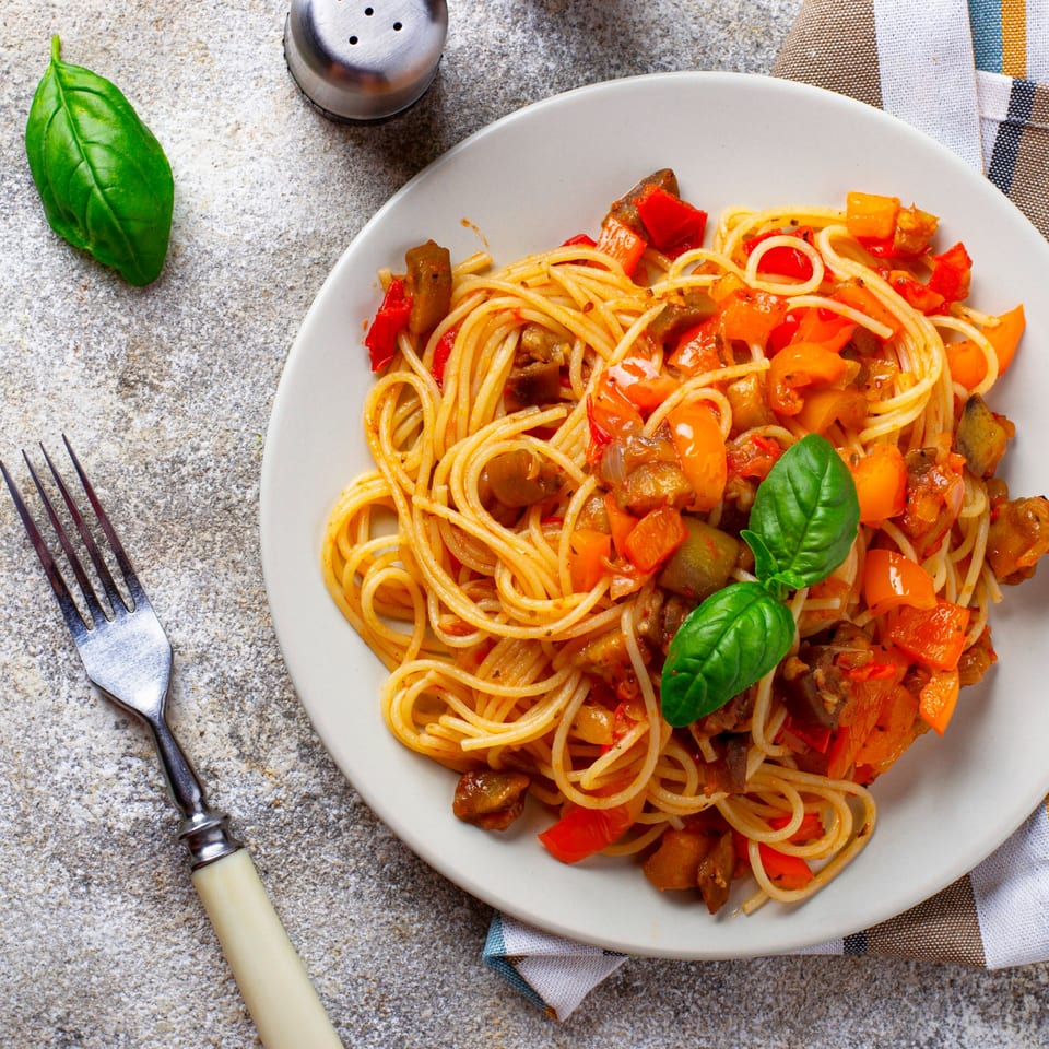 Spaghetti with colorful ratatouille vegetables and fresh basil on a white plate with a fork.
